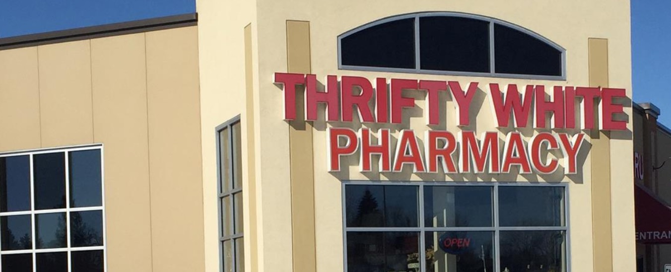 The front of a Thrifty White Drug Pharmacy building with large red letters spelling the store name above the entrance. A lit "OPEN" sign in the window indicates the store is open for customers.