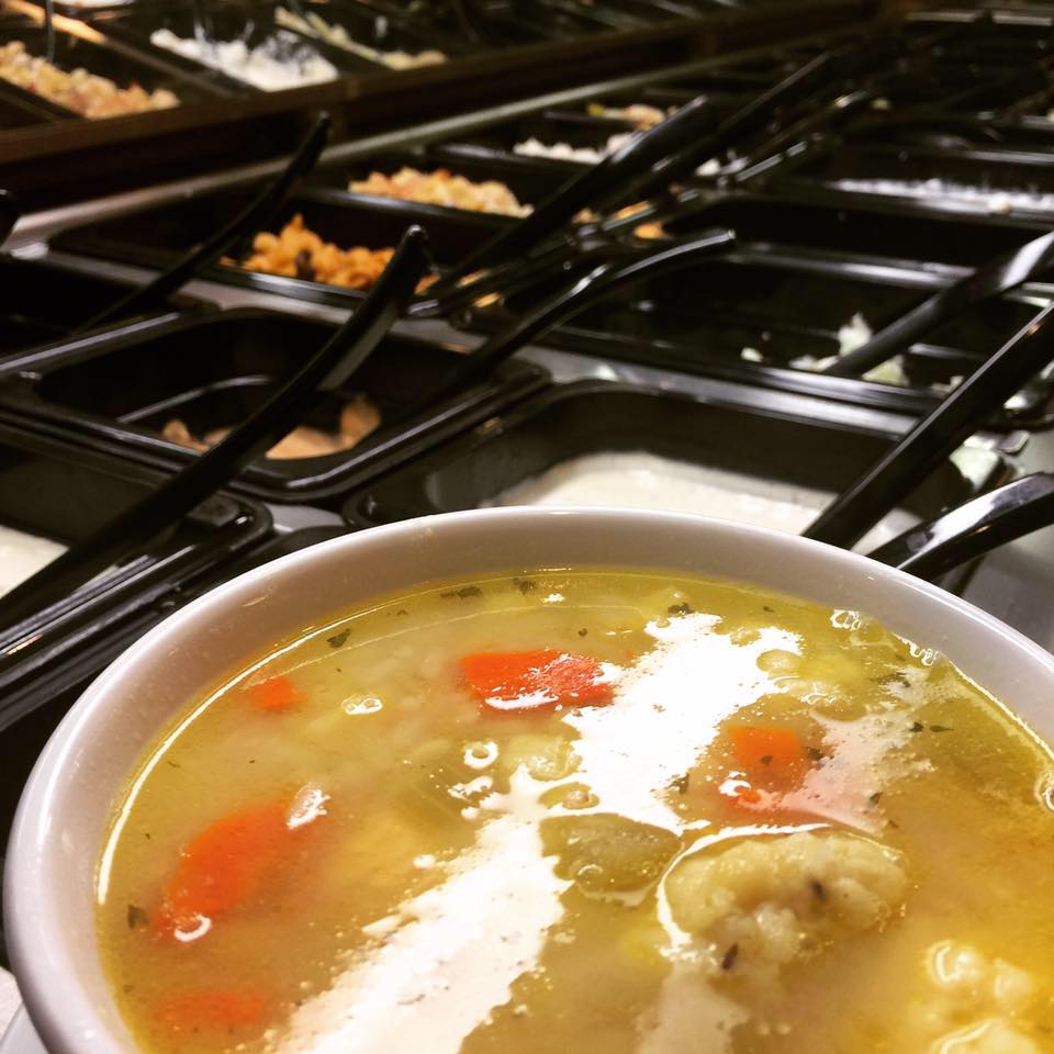 A close-up of a bowl of vegetable soup with carrots and potatoes, served at Traveler's Inn Cafe, sits in front of a self-serve salad bar featuring various toppings and dressings in black containers.