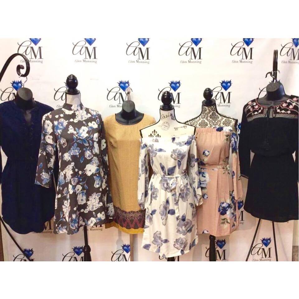Six mannequins display a variety of women's dresses in floral and solid patterns, arranged in front of a white backdrop with blue and black "CM Glen Manning" logos, embodying the latest trend and couture styles.