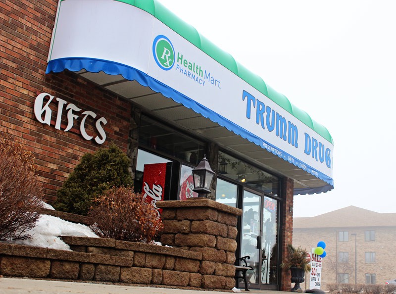 A brick building with a blue and green awning that reads "TRUMM DRUG" and "Health Mart Pharmacy." The Trumm Drug entrance displays "GIFTS," with snow on the ground and shrubs near the door.