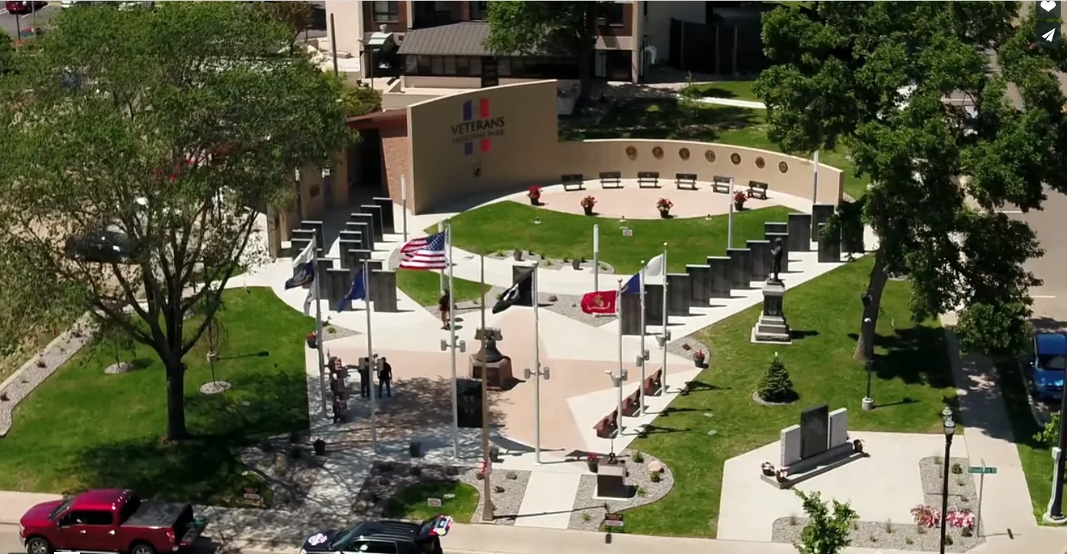 Aerial view of Veterans Memorial Park featuring a curved wall with emblems, multiple flagpoles with flags, a statue, paved walkways, green grass, benches, and trees surrounding the area.