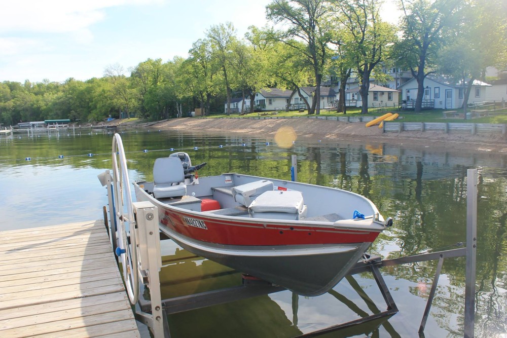 A small red and gray motorboat, available through rentals at Viking Bay Resort, is docked at a wooden pier on a calm lake, with houses, trees, and a sandy beach with yellow slides in the background.