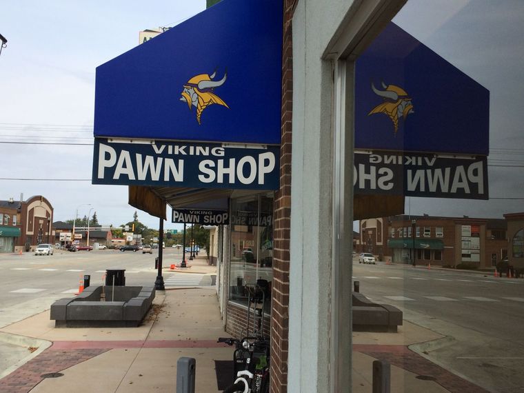 A blue awning with a Viking logo and the words "Viking Pawn Shop" covers the quiet sidewalk outside the Viking Pawn Shop, its large window reflecting the storefront and nearly empty street.