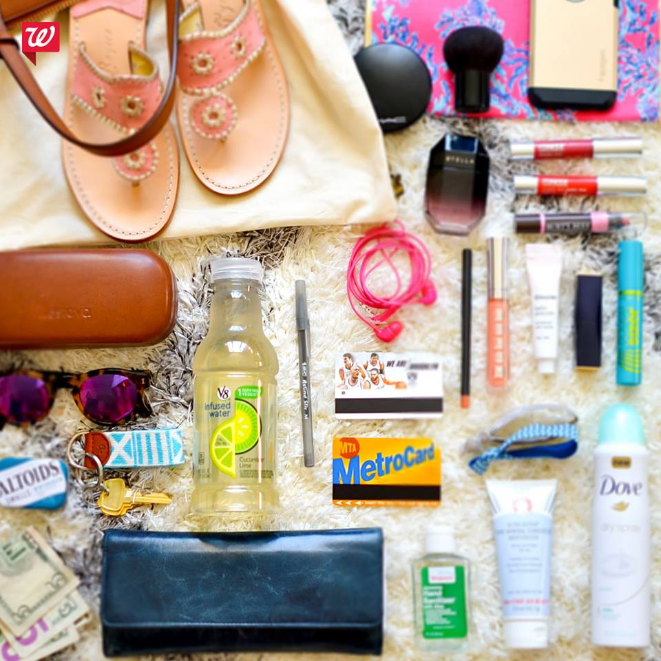A variety of items laid out, including sandals, sunglasses, cash, a wallet, keys, a MetroCard, lipstick from Walgreens, skincare products, headphones, a drink, mints, and a phone, all arranged on a flat surface.