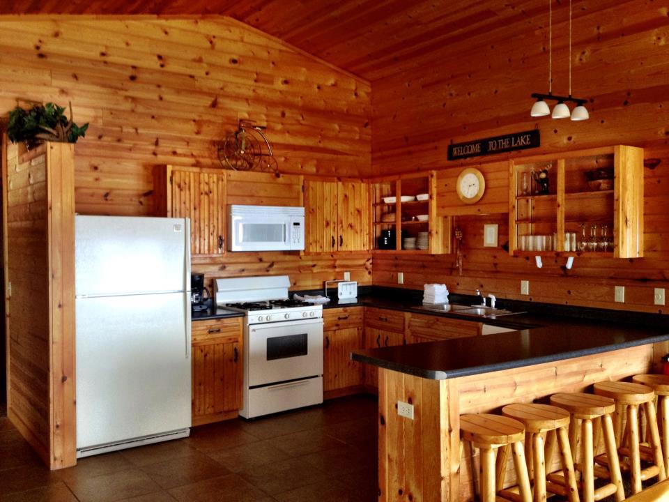 A cozy kitchen at Woodland Resort with wooden walls, cabinets, and ceiling features a white refrigerator, stove, and microwave. There’s a counter with four wooden stools, shelves with dishes and glasses, and a “Welcome to the Lake” sign above.