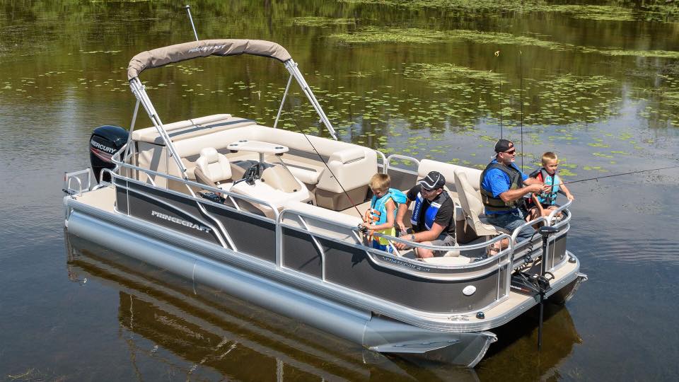 A family of four, all wearing life jackets, enjoys fishing from a pontoon boat on a calm lake surrounded by greenery and lily pads. Relaxing on the deck, they make the most of their day with Rentals at Woodland Resort.