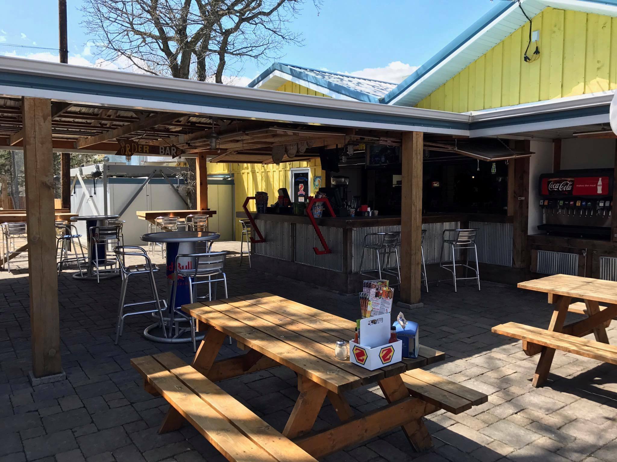 Outdoor bar and seating area with wooden picnic tables, metal stools, a Zorbaz Red Bull table, and a Coca-Cola soda dispenser. The bar sits under a wooden roof with bright yellow walls in the background on a sunny day.