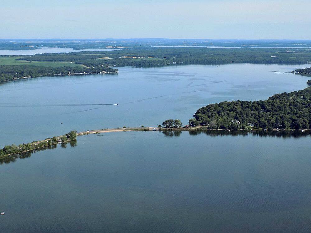 Aerial tour of Alexandria, Minnesota reveals a calm, expansive lake bordered by dense green forest and a narrow strip of land extending into the water under a clear sky.