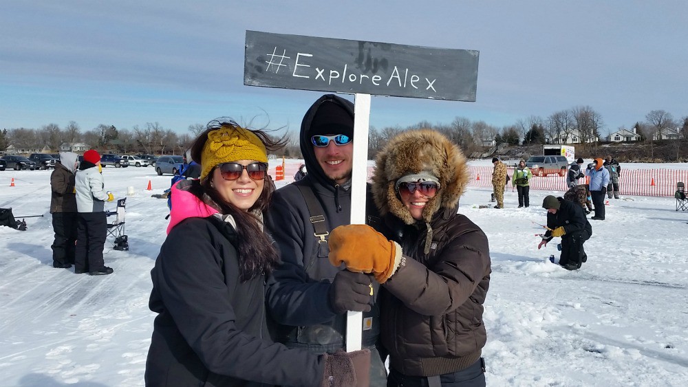 Three people dressed in winter clothes stand on snow holding a sign that reads "#ExploreAlex" near an ice fishing event in Alexandria, MN. Other people are visible in the snowy background, along with orange cones and a fence.