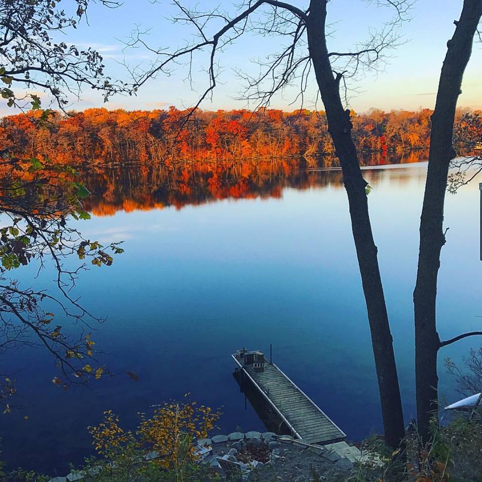 A wooden dock extends into a calm lake, reflecting autumn trees in shades of orange and yellow—a scene perfect for a collection of your fall color photo submissions. Bare branches frame the view under a clear blue sky.