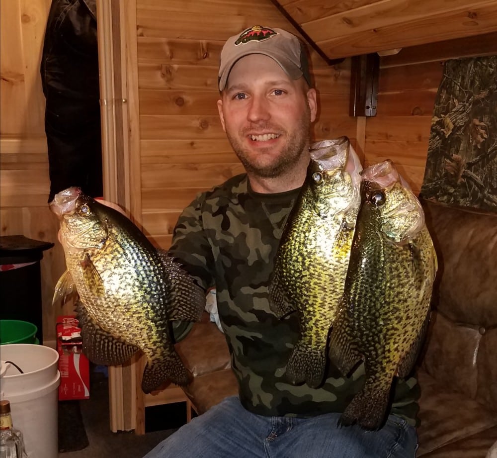 A smiling man in a camouflage shirt and baseball cap sits indoors, proudly holding up four large crappies—two in each hand. The room’s wooden walls create a cozy, cabin-like atmosphere.