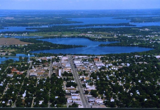Aerial view of Alexandria Aviation’s small city with a grid layout, surrounded by green trees and several blue lakes in the background under a clear sky.
