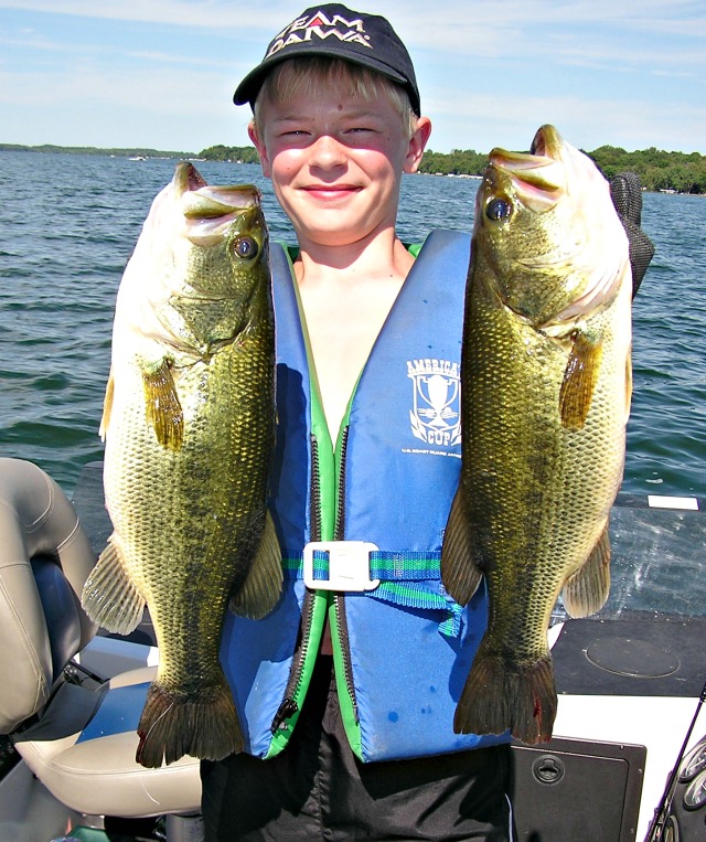 A smiling boy in a life vest and hat stands on a boat, proudly holding up two large fish—proof that Fishing Techniques For Minnesota Anglers really pay off—with a lake and trees visible in the background.
