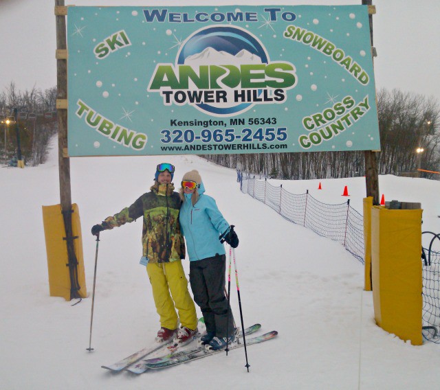 Two people in winter ski gear stand on snow with skis, posing before a "Welcome to Andes Tower Hills" sign at a winter sports area, surrounded by snowy trees and fencing.