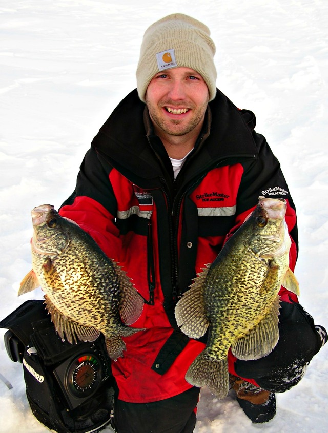 A smiling person in a red and black winter jacket and beige beanie holds up two large crappie fish, kneeling on snow-covered ground after ice fishing—showcasing effective late season crappie tactics.