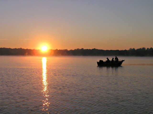 A small boat with several people is silhouetted against the water at sunrise, the sun low on the horizon and its reflection shimmering across a calm lake. Trees line the distant shore in this peaceful September 2014 scene from an Alexandria Fishing Report.