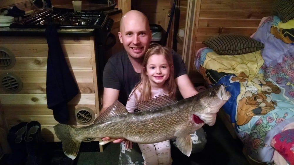 A smiling man and young girl pose indoors, proudly holding a large fish they caught while ice fishing. They are in a cozy wooden room with a bed featuring cartoon character sheets in the background.