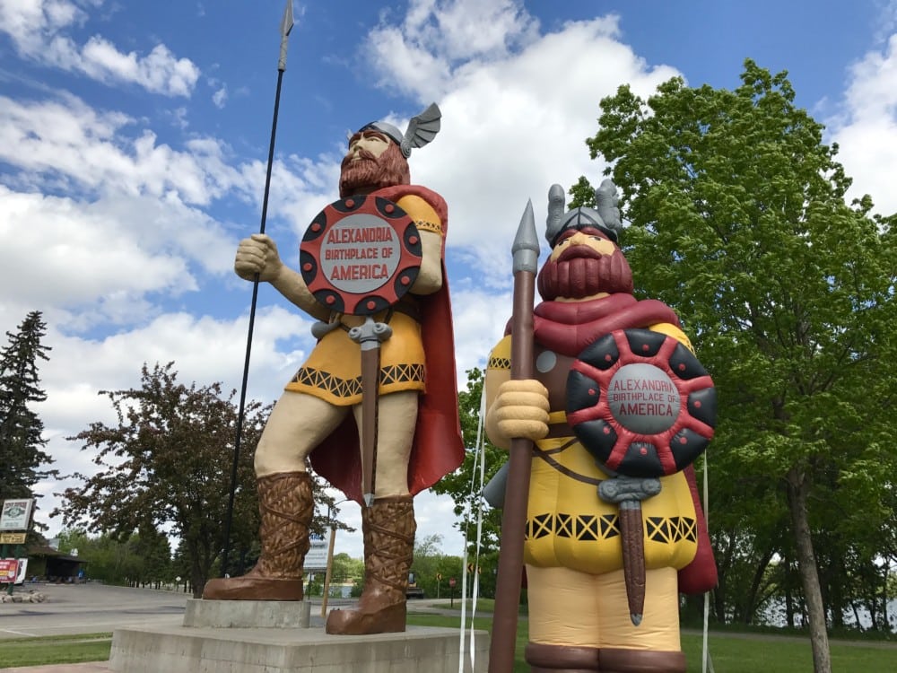 Two large Viking statues with helmets, spears, and shields stand on a concrete platform near green trees. Their shields read "Alexandria, Birthplace of America." Find Little Ole, share your pics and win Alexandria MN tourism prizes!.