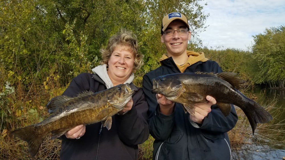 Two people smiling and holding large fish in their hands, standing outdoors by a river or lake with green trees and bushes in the background—a great fishing update at the end of October.