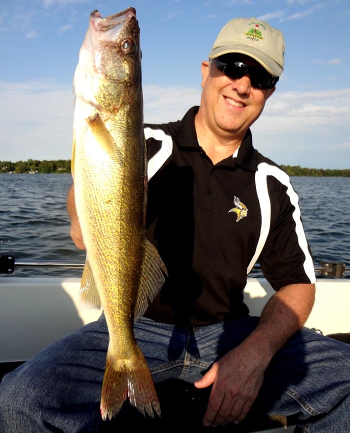 Smiling man wearing sunglasses and a cap, sitting in a boat and proudly holding up a large walleye with one hand. Water and distant trees are visible in the background under a blue sky.