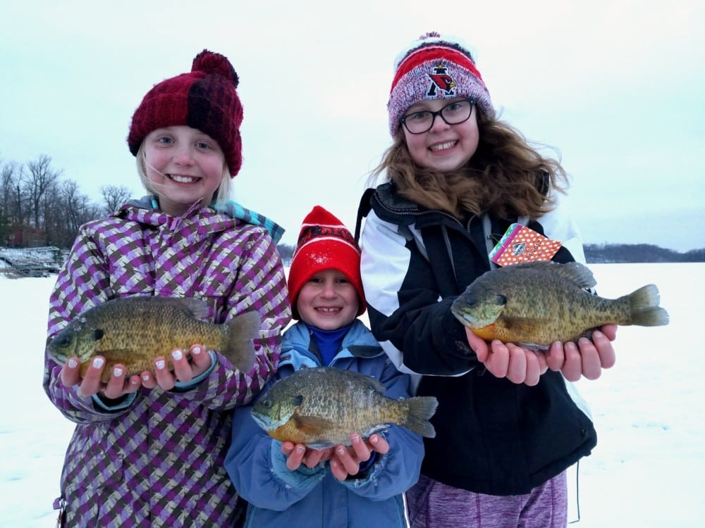 Three smiling children dressed in winter clothing stand on snow holding large fish they caught, with trees and a frozen lake featuring fishable ice in the background.