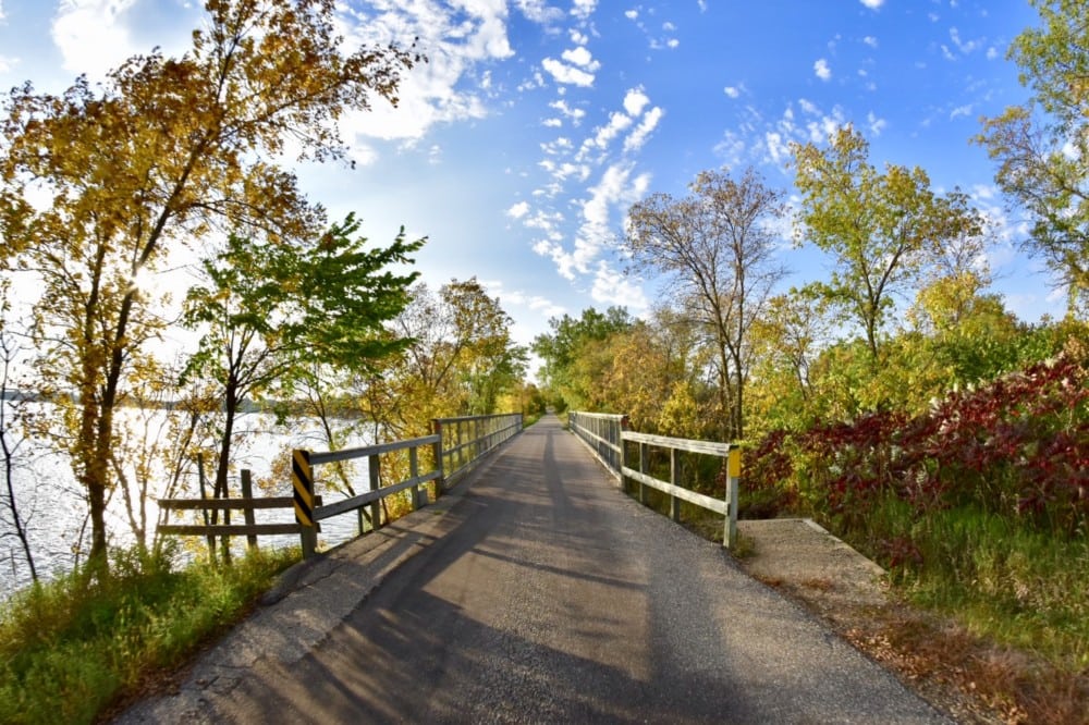 A paved trail with wooden railings winds between trees, their fall colors progressing beautifully in Alexandria Lakes Area, beside a calm body of water under a partly cloudy blue sky.