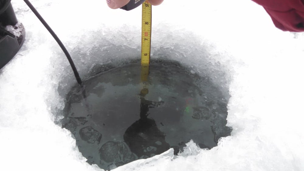 A hand holds a measuring tape in a hole drilled through thick ice on a frozen surface, checking the depth of the water—a typical scene for an Ice Update on current lake conditions in Alexandria Area Lakes.