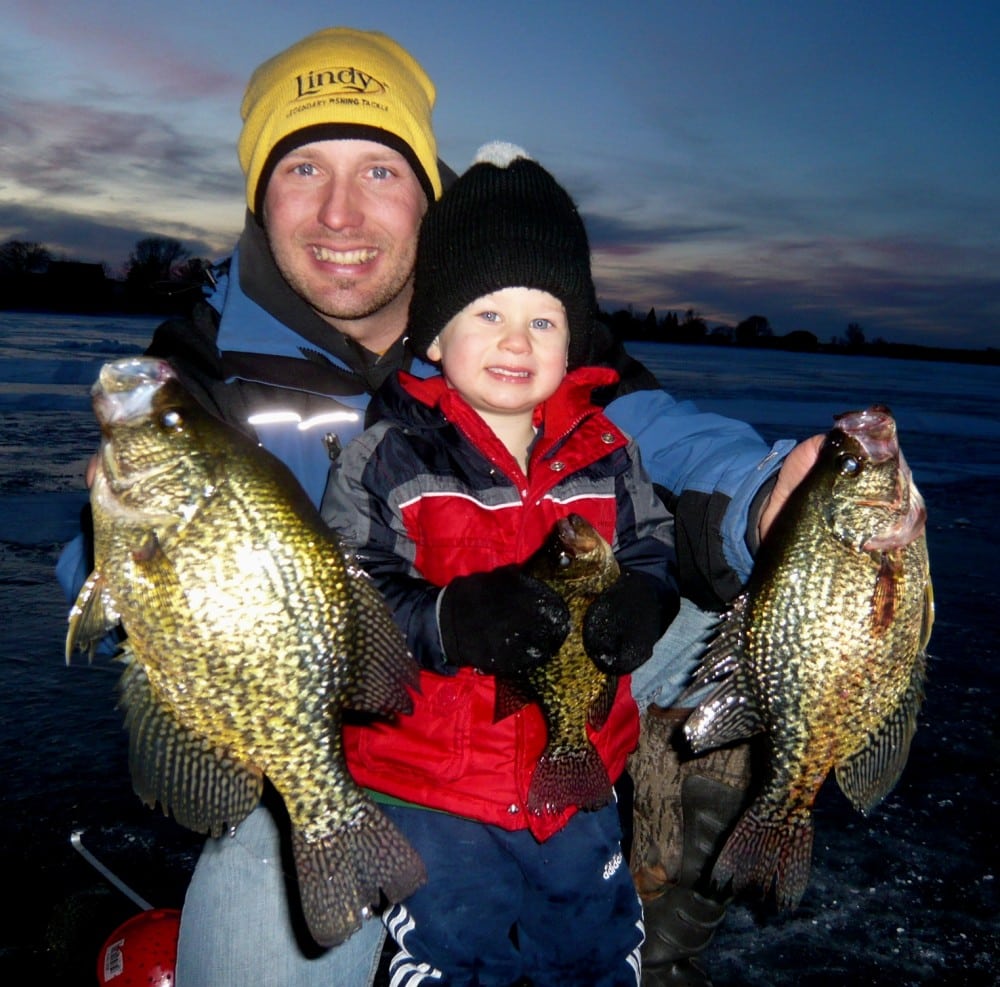 A smiling man and young child, both bundled in winter clothing, proudly hold up three large crappies while crappie fishing on a frozen lake near Alexandria at dusk.