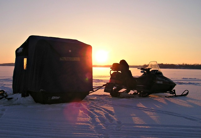 A snowmobile is parked on a snowy landscape at sunset, towing a dark-colored ice fishing shelter—highlighting the excellent ice fishing conditions in Alexandria as long shadows stretch across the gleaming snow.