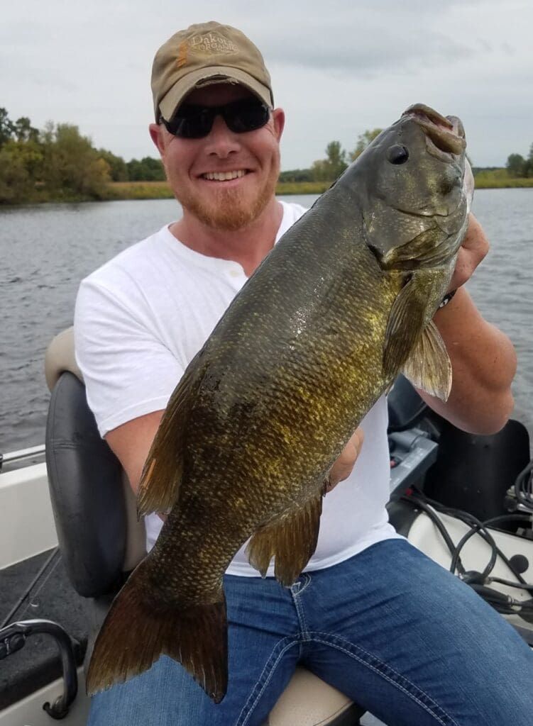 A smiling man wearing sunglasses and a cap sits in a boat on Alexandria Lakes, proudly holding up a large Smallmouth Bass he caught. Trees and cloudy skies are visible in the background.
