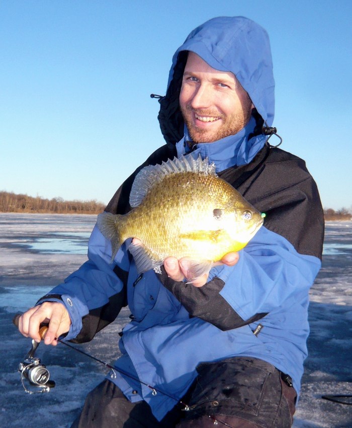 A person in a blue jacket smiles while holding a big panfish in Alexandria, sitting on an icy surface with a fishing rod in hand under a clear blue sky.