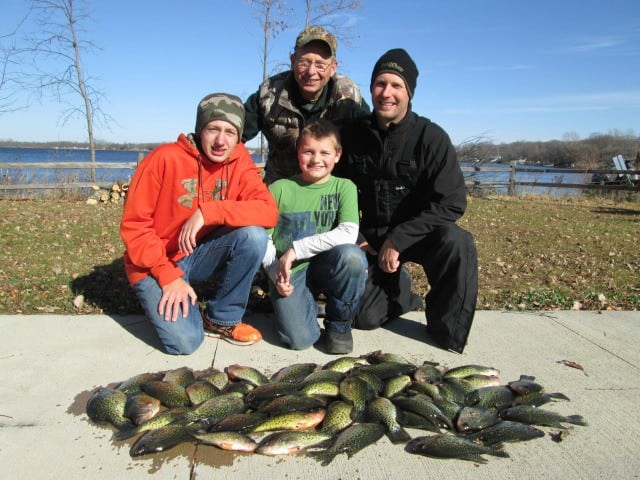 Four people posing and smiling outdoors near a lake, crouched beside a large catch of fish laid out on the ground. It’s a sunny day with clear skies and trees in the background, celebrating their haul from one of the season's First Frozen Lakes.
