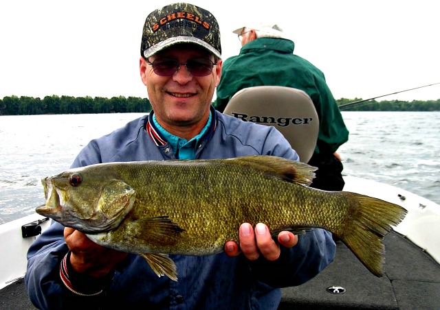 A man wearing glasses and a cap smiles while holding a large fish on a boat, with another person in the background. The scenic lake and trees set the scene for this memorable catch, perfect for any June Lake fishing report.