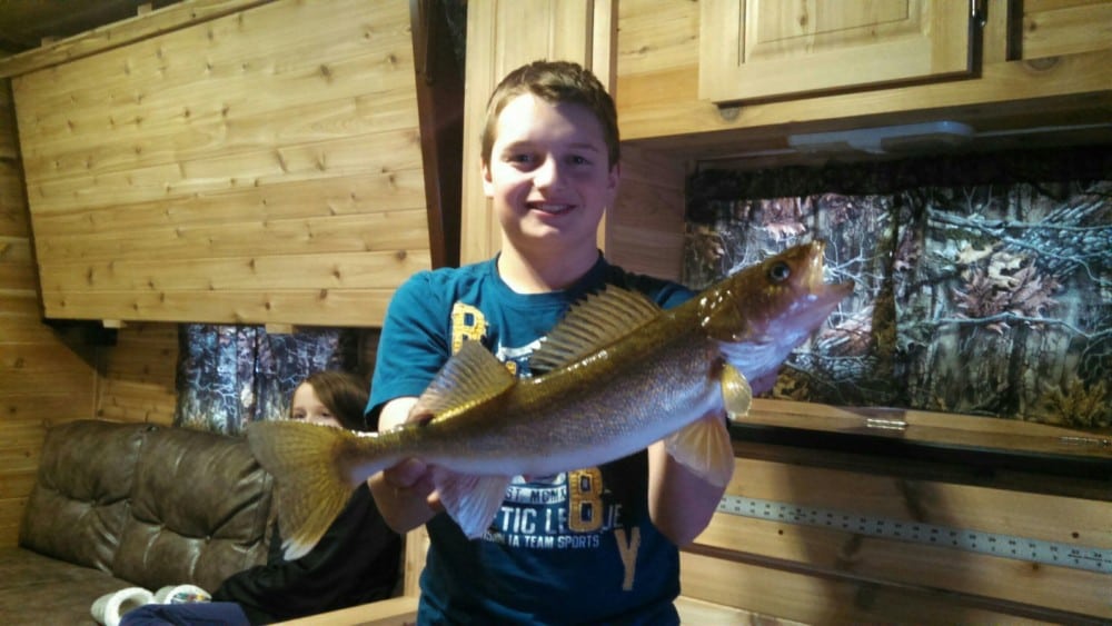 A smiling boy in a blue shirt proudly holds his catch for big fish pictures inside a wood-paneled room, possibly a cabin, while another person relaxes on a couch in the background.