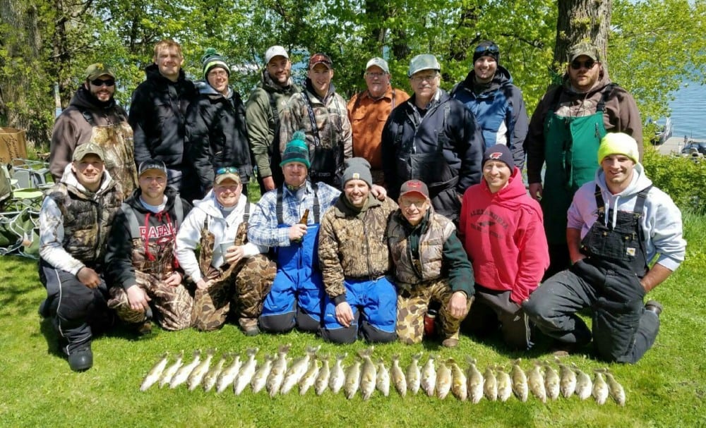 Seventeen people in outdoor clothing pose in two rows on grass, behind a display of freshly caught fish laid out in a row—proof that the 2016 walleye season is off to a great start near Alexandria, MN. Trees and lakeside scenery complete the backdrop.