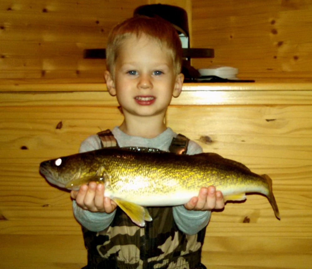 A young boy in camouflage overalls smiles while holding a large crappie with both hands in front of a wooden wall indoors.