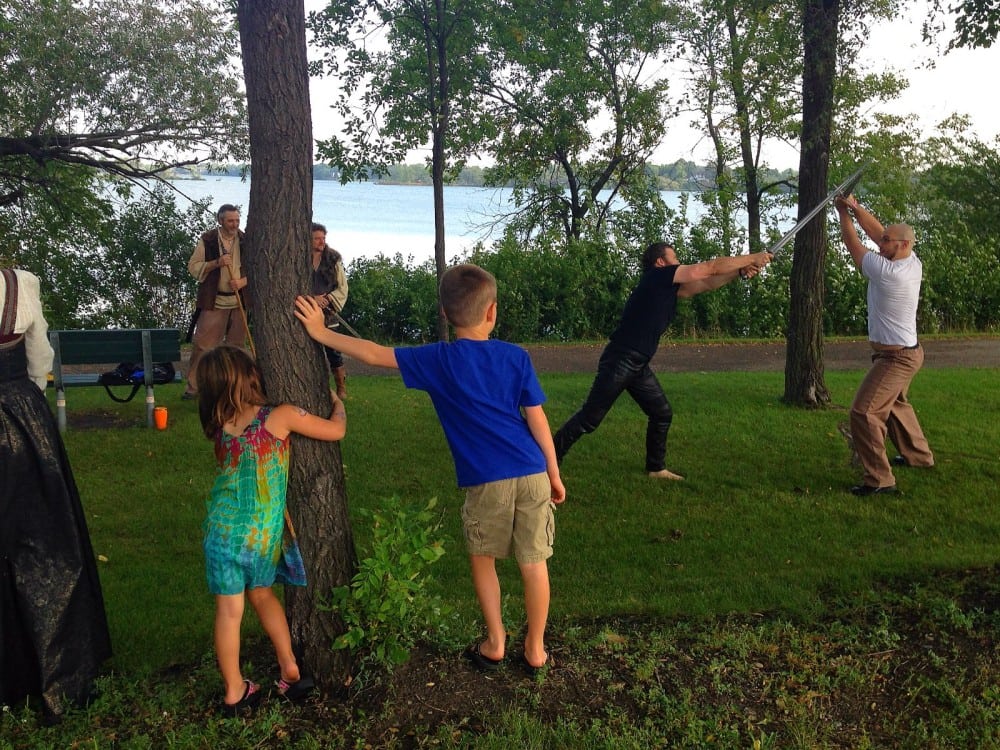 Two kids watch as two adults have a dramatic sword fight in a park near a lake, while others in costumes—suggesting a Viking adaptation or medieval fantasy event—stand nearby, reminiscent of Hamlet performed at the Alexandria Farmers Market.