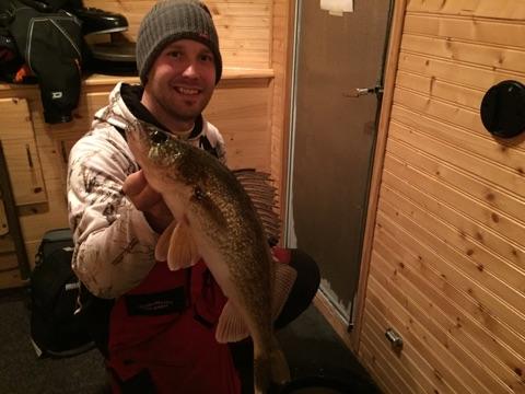 A smiling person in winter clothing holds up a large fish inside a wooden-paneled room, likely an ice fishing hut—proof of excellent fishing and great ice thickness, making for a successful day on the ice.