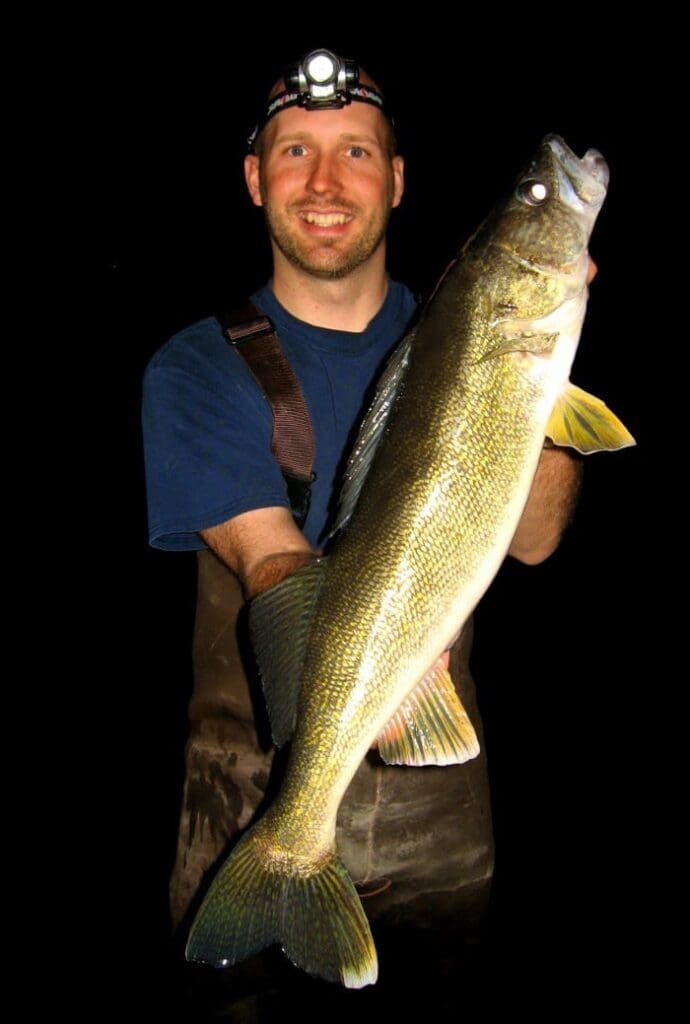 A smiling man wearing a headlamp and waders holds a large fish, likely caught at night during the 2015 Walleye Opener, against a completely black background.