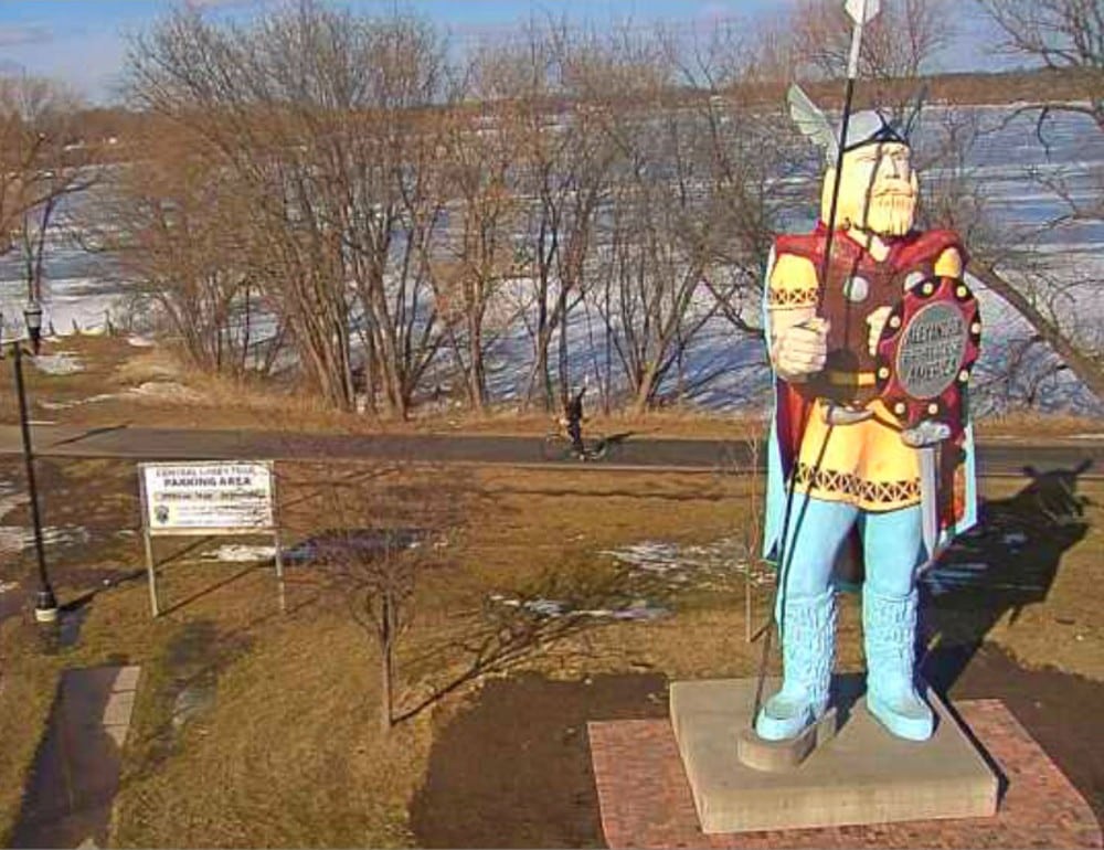 A large Viking statue holding a spear and shield stands on a brick base near the walking path, offering a unique landmark for those checking central lakes trail conditions. Patchy snow covers the ground, with bare trees under a clear sky in the background.
