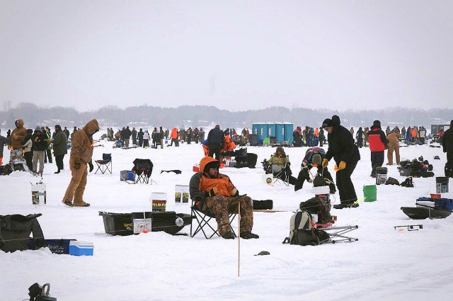 Hundreds of people bundled in winter clothing are taking on a fishing challenge, ice fishing on a frozen lake surrounded by gear, chairs, and portable shelters under a cloudy sky.