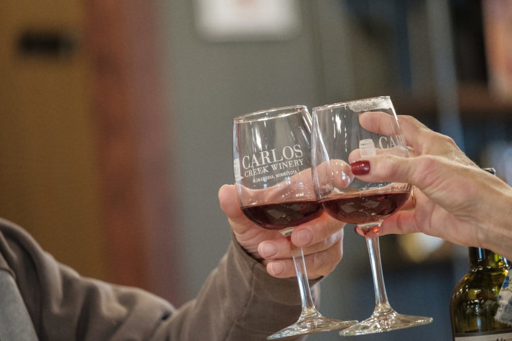 Two people clinking "Carlos Creek Winery" red wine glasses, celebrating a romantic getaway indoors—a perfect Valentine's Day moment.