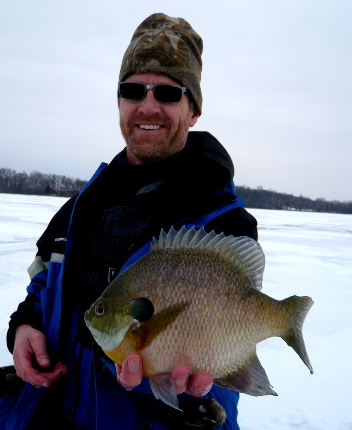 A man wearing sunglasses and a camouflage hat smiles while holding a trophy bluegill in front of him on a frozen, snowy lake. Trees are visible in the background under a cloudy sky.