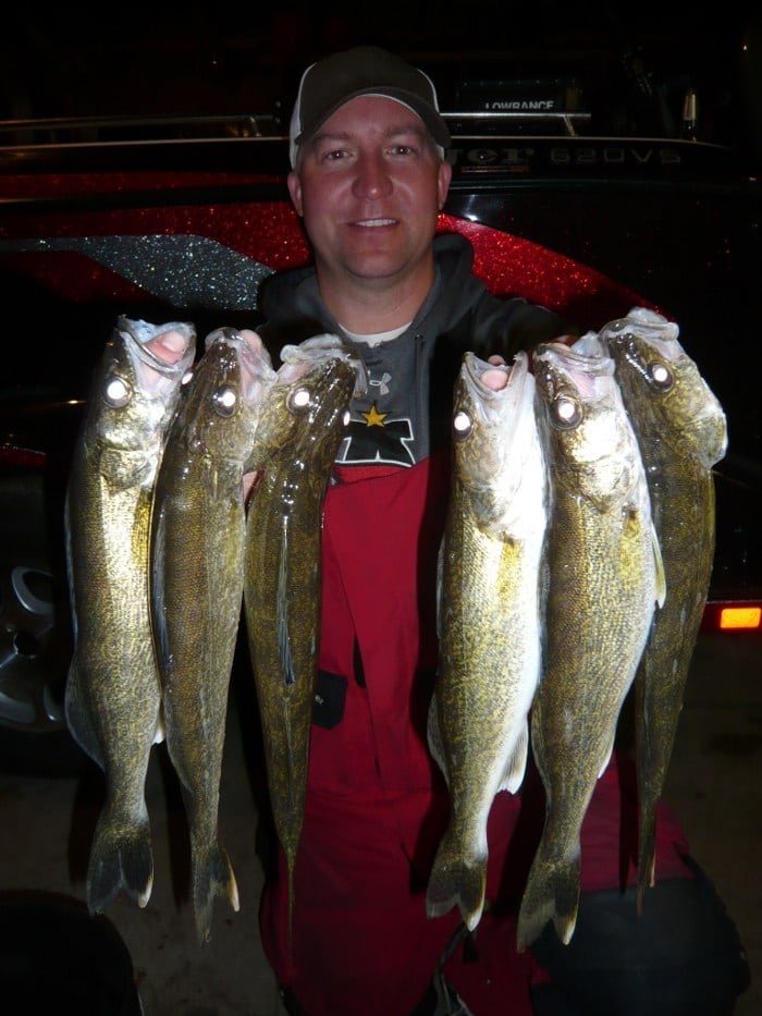 A smiling person in a cap and red jacket holds up six large fish, likely from a successful day out. The scene suggests a memorable catch, perfect for a May 2015 walleye fishing report, with the background showing a boat or vehicle.