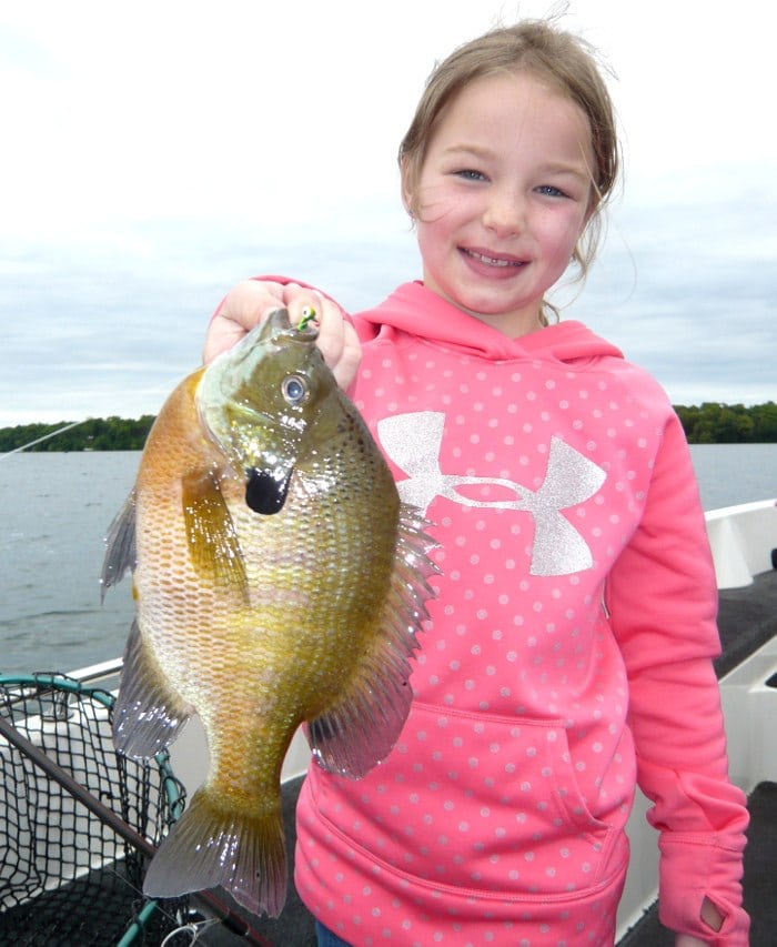 Smiling young girl in a pink hoodie holds a large fish with one hand while standing on a boat, water and trees in the background—perfect for this year’s 4th of July fishing report.