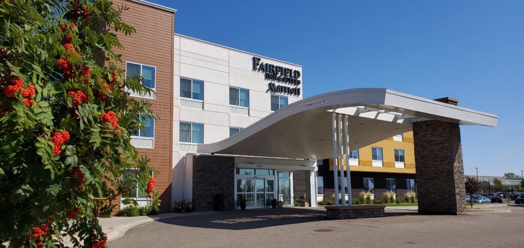 A modern Fairfield Inn & Suites Alexandria hotel entrance with a curved white canopy, stone pillars, large glass doors, and a tree with clusters of red berries in the foreground under a clear blue sky.