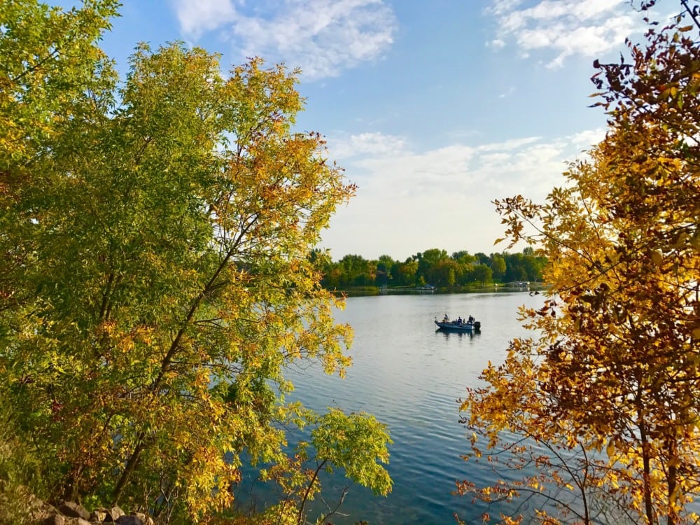 A small boat with people floats on a calm lake surrounded by trees, showing green and golden hues in this med September fall color update, beneath a partly cloudy blue sky.