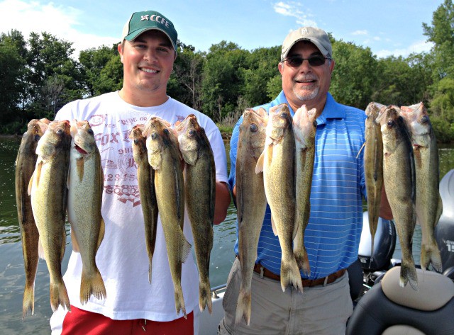 Two men standing outdoors on a boat in Alexandria, each holding up several large fish they caught, smiling at the camera. Trees and water are visible in the background, capturing a great moment from the August Fishing Report.