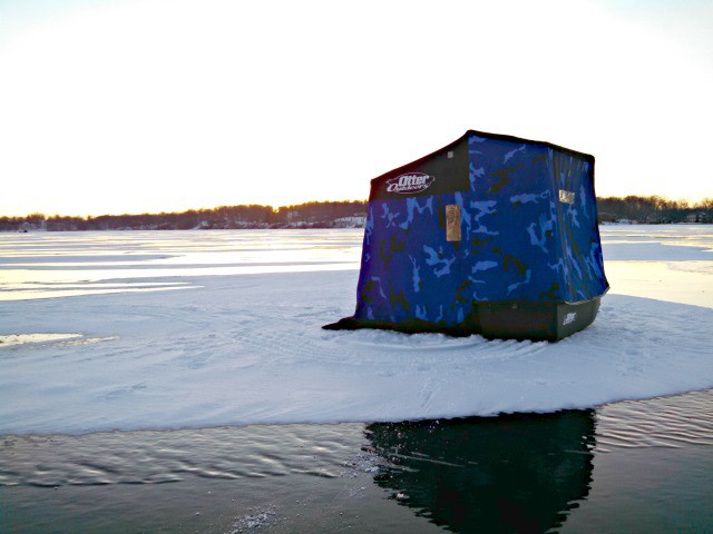 A blue ice fishing shelter sits on a snowy, frozen lake near an open area of water under a bright sky at sunset. Trees line the distant shore, creating a peaceful scene for fishing enthusiasts.