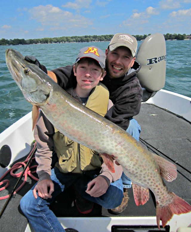 Two people sitting on a boat, smiling and holding a very large fish—likely a muskie—caught using mid-summer fishing tactics, with a lake and trees in the background under a partly cloudy sky.